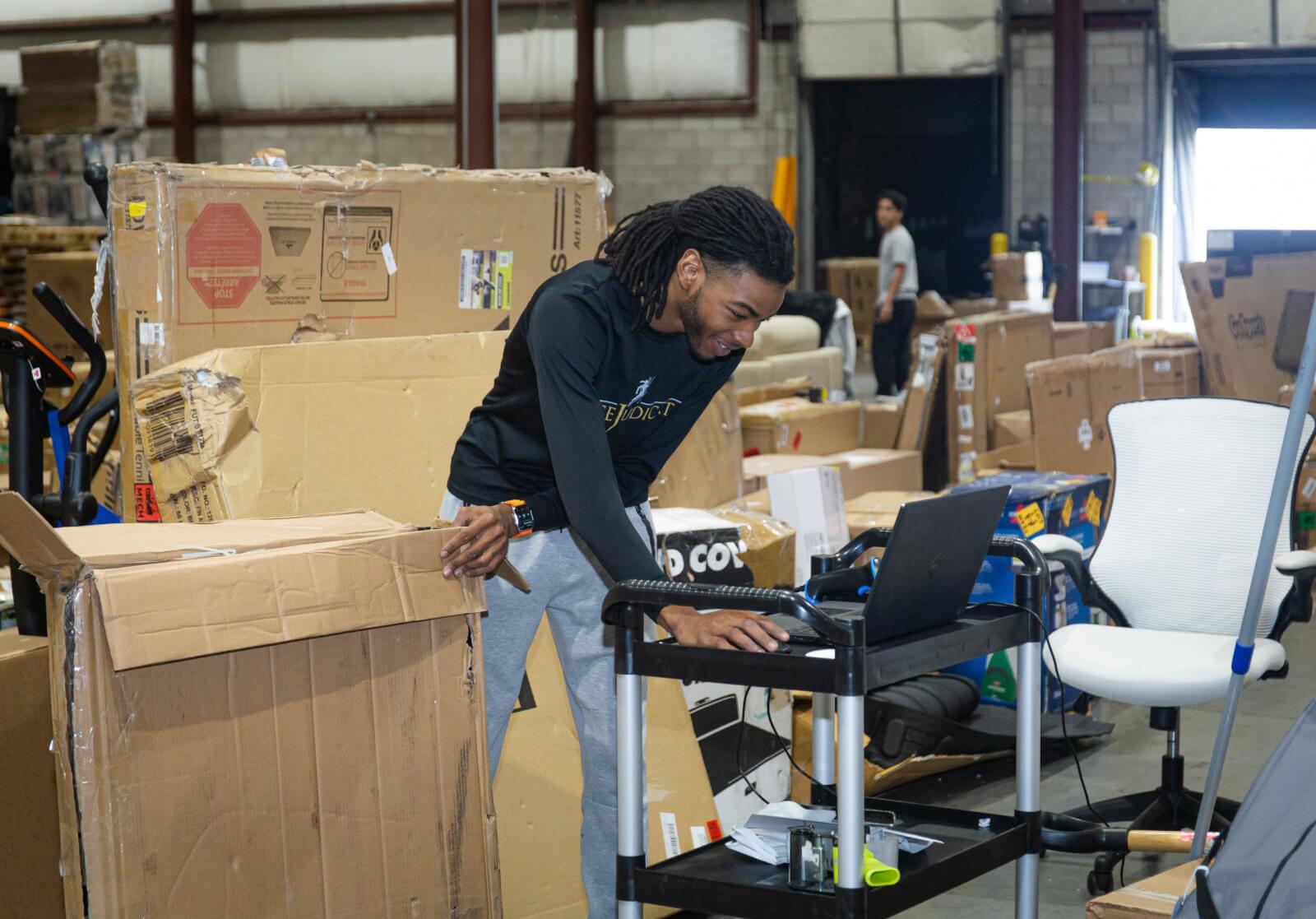 Man typing on a computer in a warehouse filled with cardboard boxes.