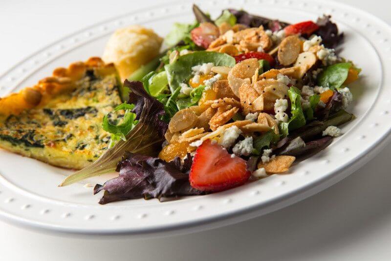 A plate of quiche is paired with Ashley Mac's most popular baby bleu salad with fresh strawberries, mandarin oranges, bleu cheese crumbles and sugared almonds, served over a bed of mixed greens with their homemade poppyseed vinaigrette. Oval plate with quiche and salad